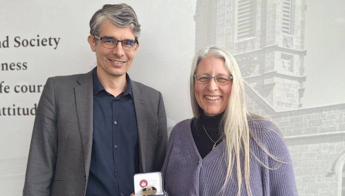 Image of a man and a woman holding a medal and standing against a wall with a greyed image.