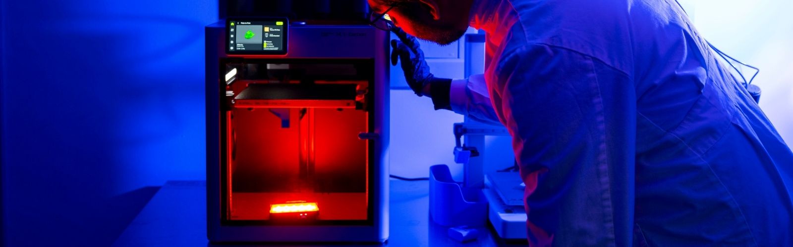 A person in a labcoat working with a3D printer. A red light is in the background