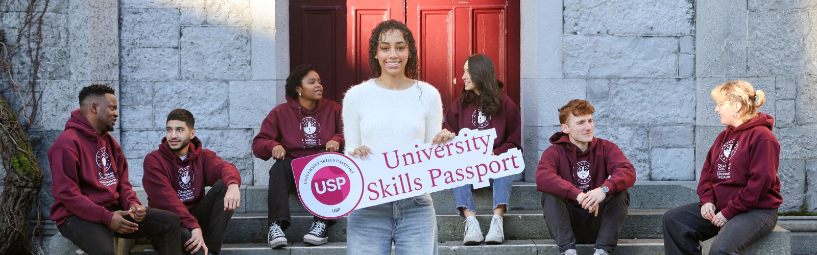 Student dressing in white top and jeans holding a USP sign, while peers sit on steps behind her in University hoodies