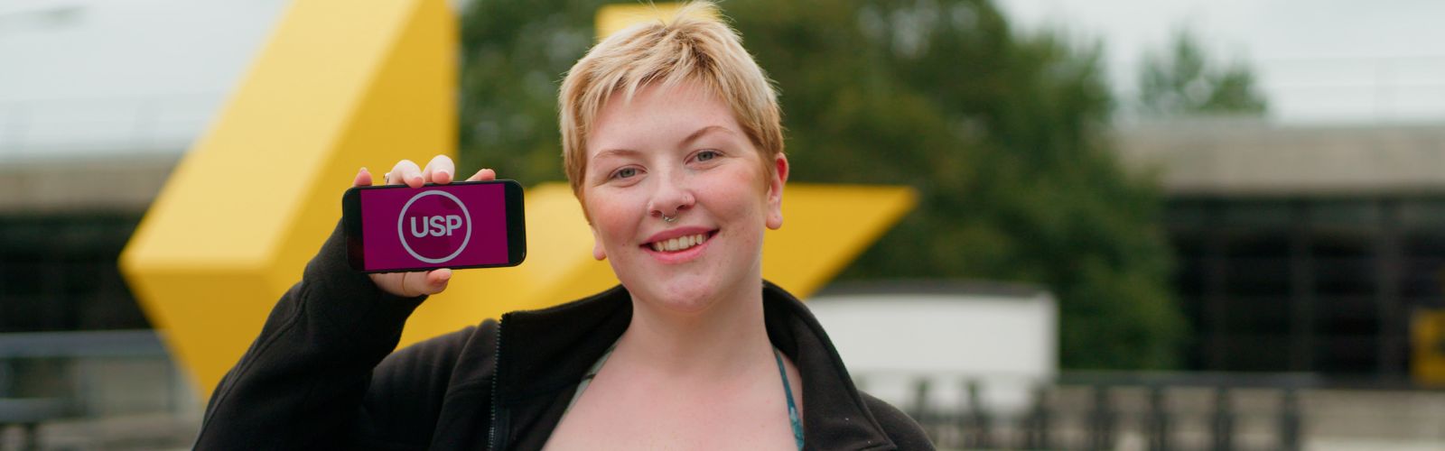 Student standing in front of the Big Yellow, looking at the camera, holding a phone with USP super imposed on it.